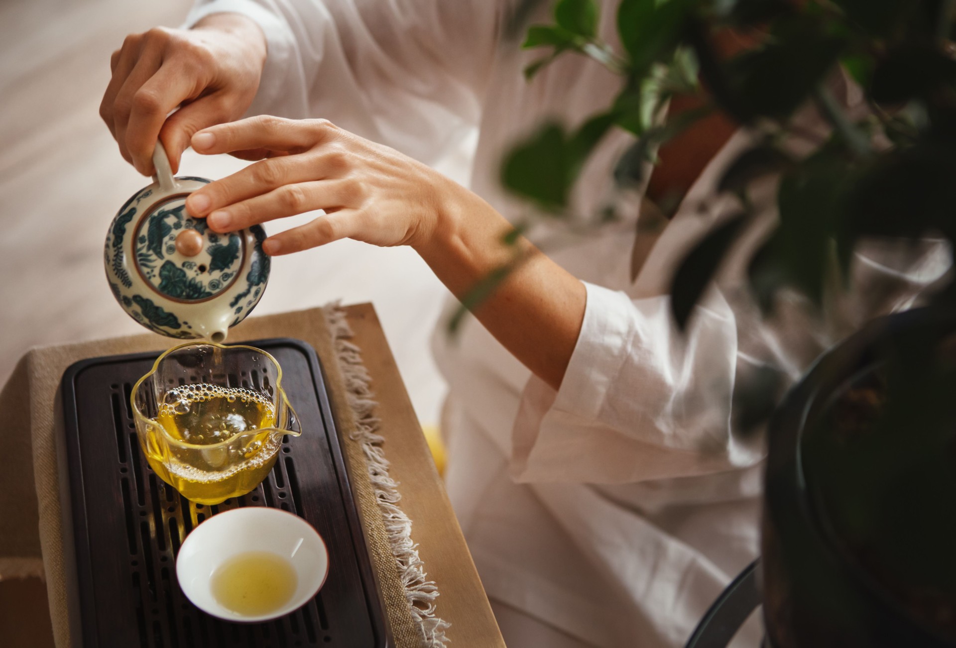 Female hands pouring tea from patterned clay teapot into clear glass pitcher, creating serene tea ritual. Soft light, wooden tea tray and greenery evoke calming mindful chenese ceremony atmosphere. Female hands pouring tea from patterned clay teapot into clear glass pitcher, creating serene tea ritual. Soft light, wooden tea tray and greenery evoke calming mindful chenese ceremony atmosphere.