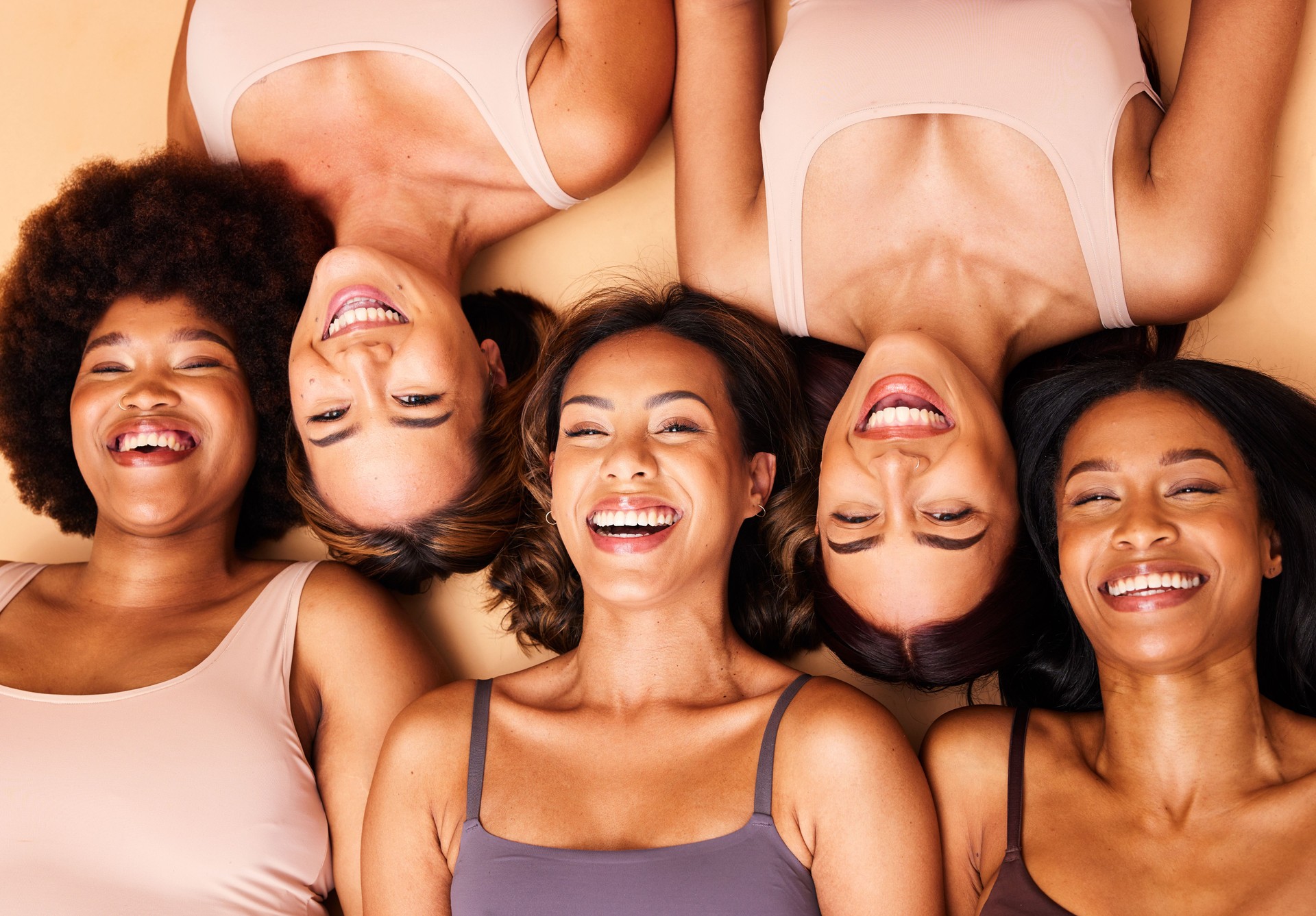 Diversity, beauty and portrait of women from above with smile, self love and solidarity in studio. Happy face, group of friends on beige background with underwear, skincare and cosmetics on floor. Diversity, beauty and portrait of women from above with smile, self love and solidarity in studio. Happy face, group of friends on beige background with underwear, skincare and cosmetics on floor.